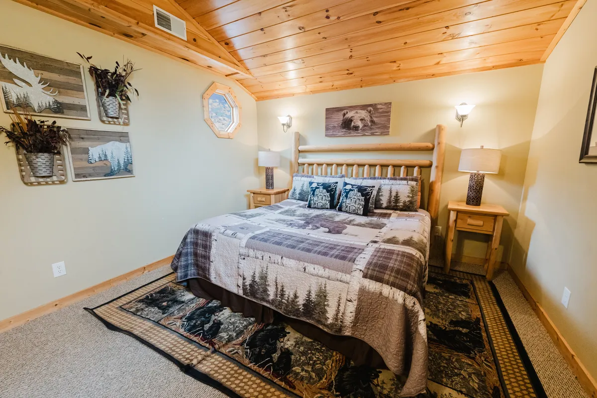 A warmly lit bedroom with a log bed, decorative pillows, nature-themed wall art, and nightstands with lamps. The inviting decor emphasizes a rustic mountain feel, and a unique octagonal window suggests a scenic view from bed.