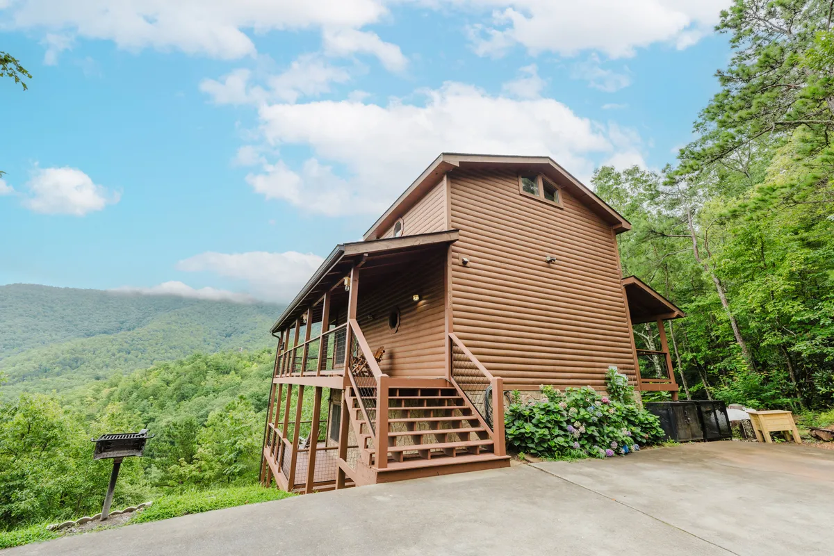 A wide exterior shot of the log cabin structure nestled among lush greenery, with expansive mountain views visible in the background and a spacious outdoor deck in the foreground. The image exudes tranquility and a sense of being immersed in nature, inviting visitors to imagine themselves enjoying the Smoky Mountains right from the property.