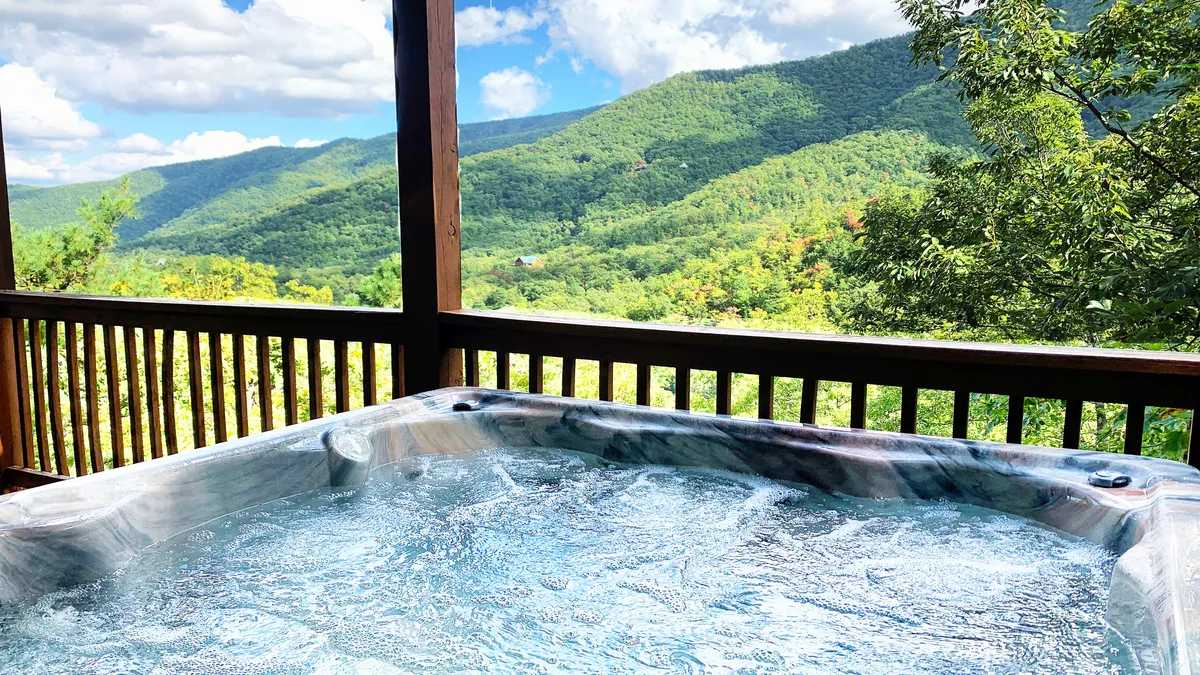 The image features the cabin's inviting outdoor hot tub perched on a wooden deck, with expansive, unobstructed views of the Smoky Mountains stretching out beyond the railing. The tranquil scene is bathed in the warm glow of sunset, highlighting the perfect spot for relaxing, enjoying morning coffee, or stargazing under the Tennessee sky.