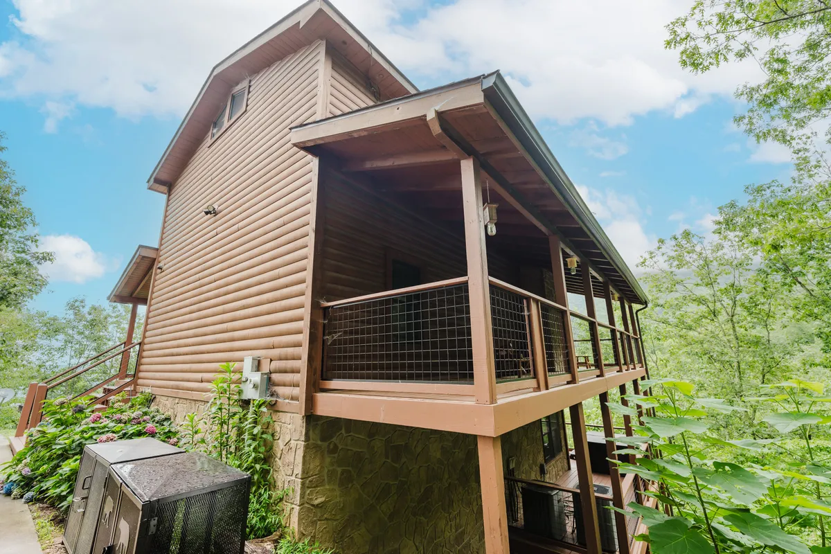 A view of the cabin's exterior featuring wooden siding, a balcony, and surrounding greenery. The space suggests privacy, adventure, and a welcoming basecamp for exploring local attractions.
