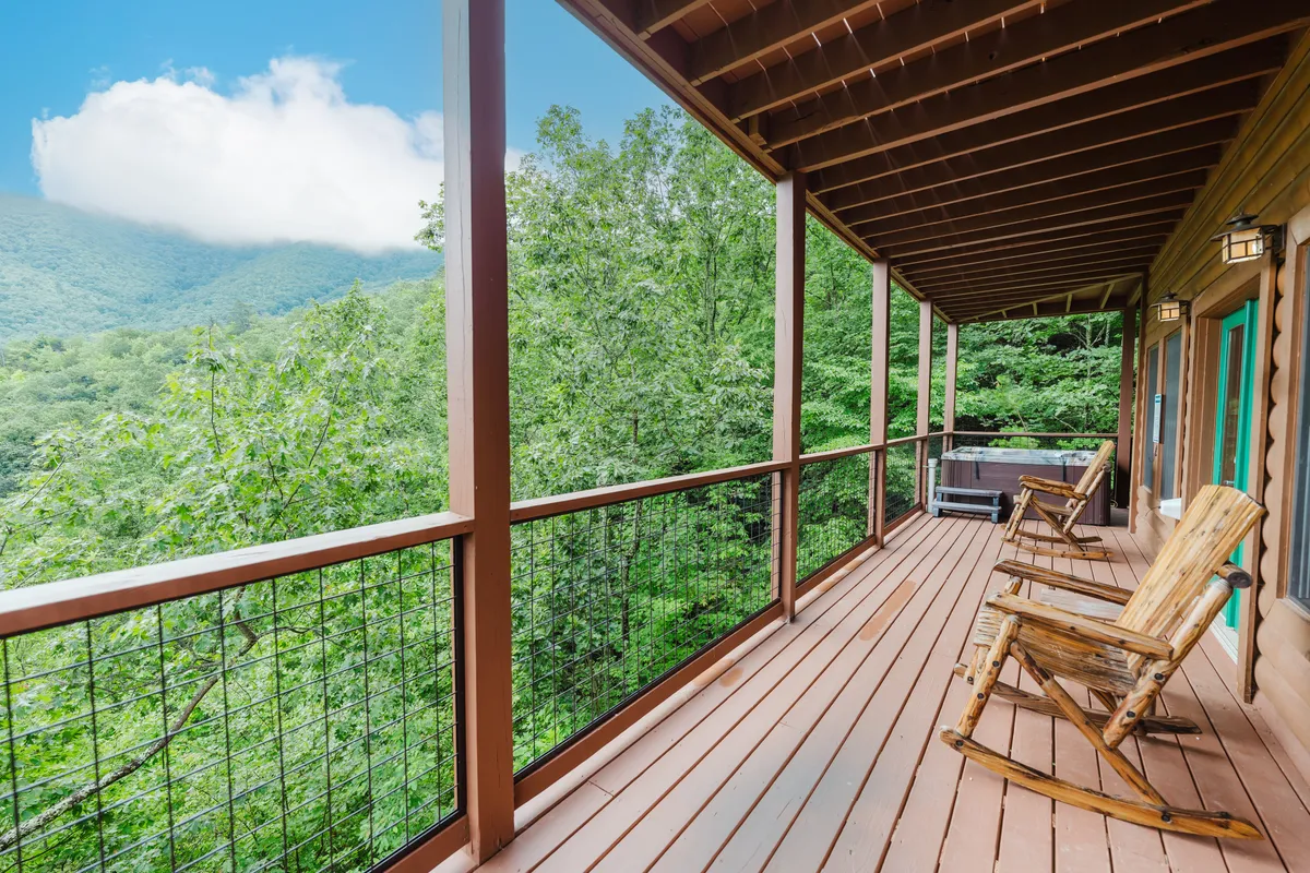 An exterior view featuring a pair of wooden rocking chairs facing out towards rolling mountains, set against a tranquil backdrop of trees and open sky. The scene is imbued with calm, highlighting the deck's role as a peaceful escape for relaxation and soaking in natural beauty.