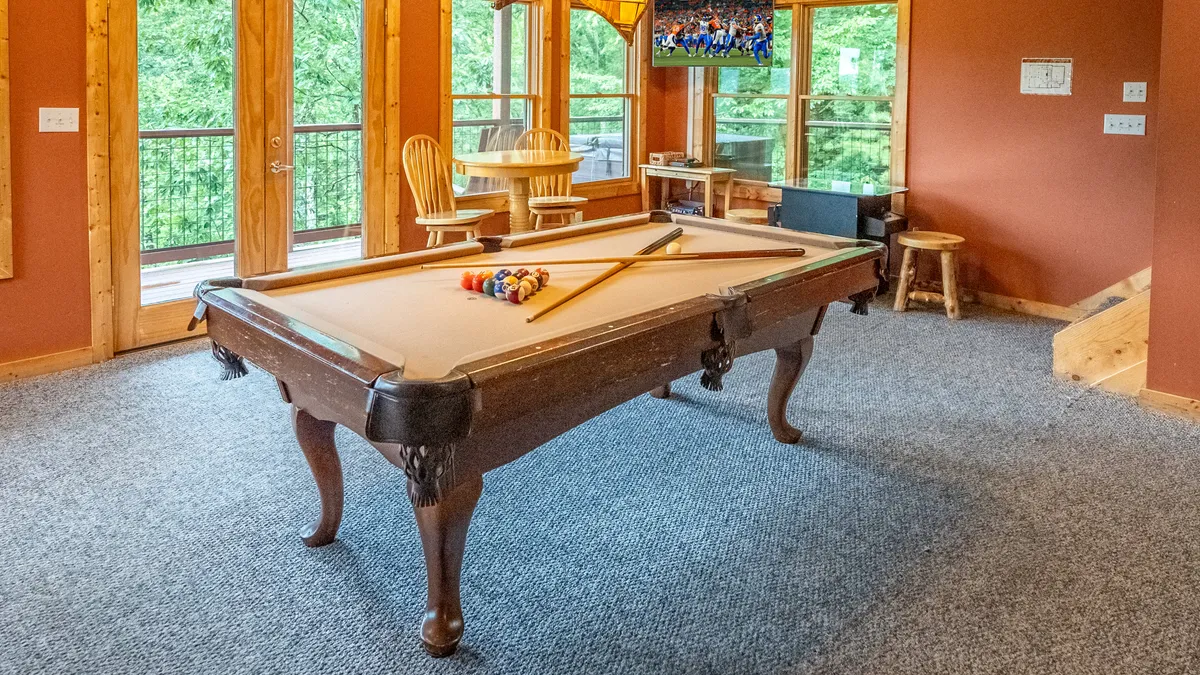 A game room designed for fun and friendly competition, featuring a pool table prominently in the center. In the background, a wall-mounted TV, a window letting in natural light, and a dining table offer multiple spots for socializing and relaxing. The scene exudes a relaxed, playful mood.