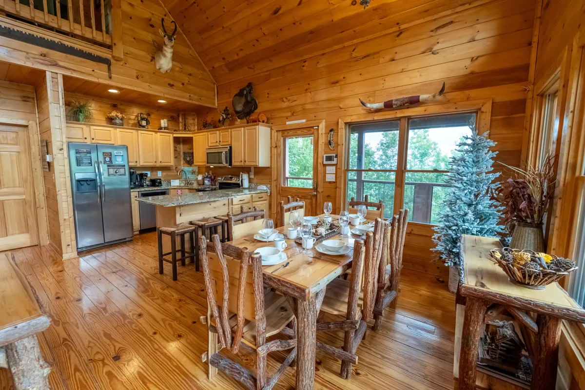A warm, open dining area showcasing a large wooden dining table adjacent to the kitchen, with large windows bringing in natural light and wooden accents throughout. The space is styled for both casual breakfasts and festive group dinners.