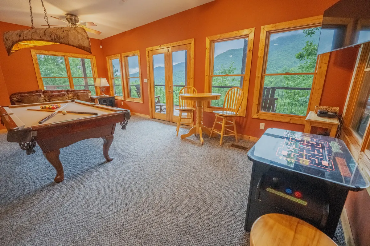 A playful and cozy game room angle featuring a pool table, a vintage arcade game, large windows with wooded views, and rustic wooden furniture. The setup invites both classic and modern gaming, with ample natural light enhancing the cheerful ambiance.