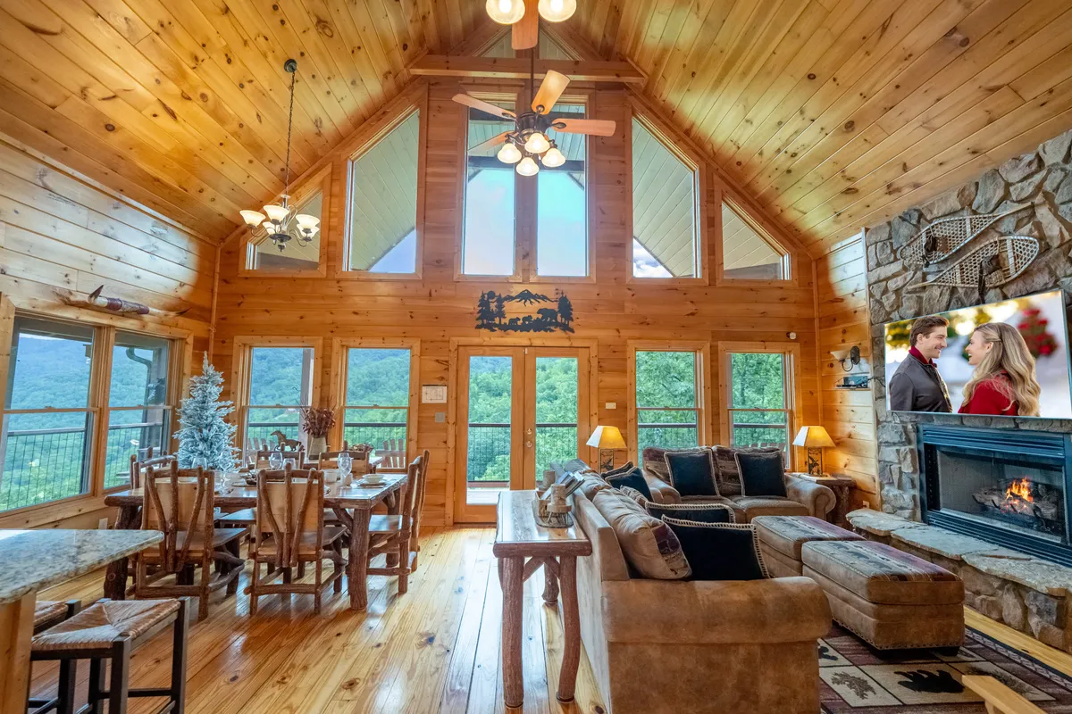 A spacious, light-filled living room with large windows that reveal mountain views. In the foreground, a plush seating area surrounds a stone gas fireplace, and a flat-screen TV is visible with a couple relaxing on screen. A wooden dining table sits nearby, enhancing the communal, inviting atmosphere.