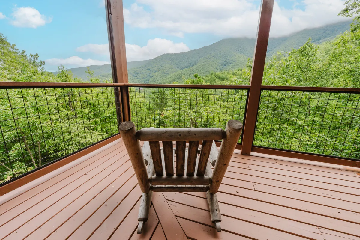 A peaceful outdoor space featuring a wooden rocking chair positioned to appreciate an expansive mountain view framed by lush greenery. The surrounding foliage suggests a vibrant, ever-changing landscape, ideal for connecting with nature and enjoying the sights and sounds of the Smokies.