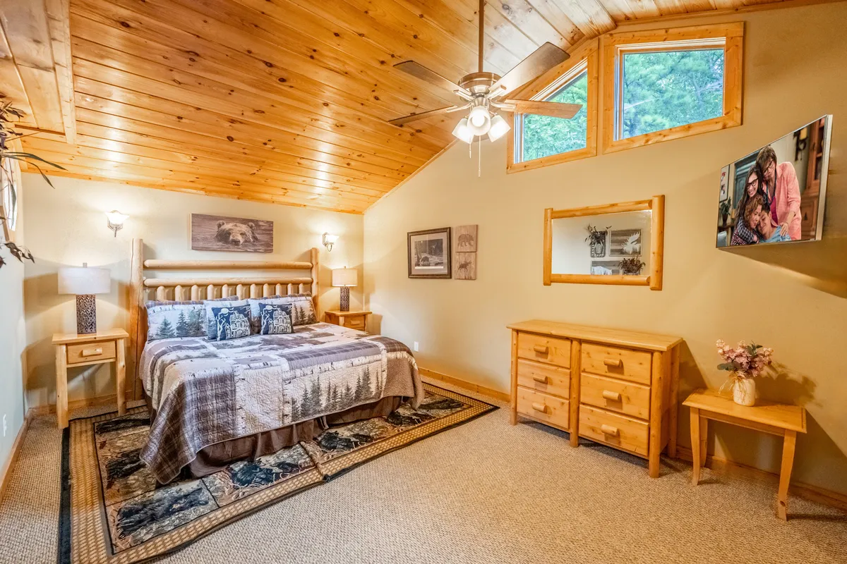 This image presents a cozy bedroom featuring a bed with patterned bedding, wooden furniture, a decorative mirror, and a wall-mounted TV. Natural light floods the space, and a ceiling fan hangs above, contributing to overall comfort.